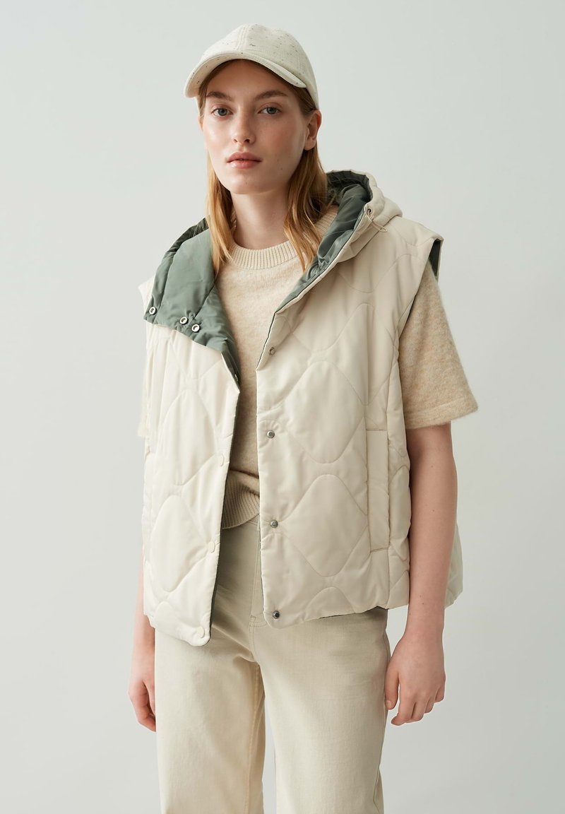 Junge Frau, die eine beigefarbene gesteppte ärmellose Jacke mit einer grünen Kapuze, ein beiges Strickoberteil, eine Hose und eine hellfarbige Mütze trägt und vor einem weißen Hintergrund steht.