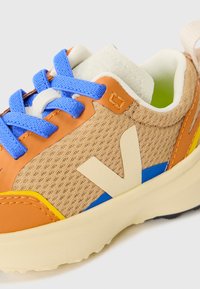 Sko med beige mesh-overdel, oransje lærdetaljer, blå snørebånd og en kremgummisåle med blå og gul design.