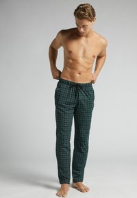 Hombre sin camiseta ajustando pantalones de pijama de cuadros en tonos verde oscuro y azul con cordón negro, de pie descalzo sobre un fondo neutro.