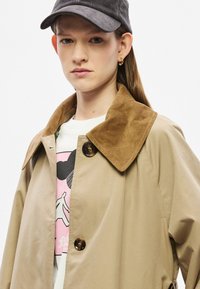 Beež värvitud trentšmantel pruuni ribikangast kraega, millel on kaks pruuni nööpi ja minimaalne läige. All on valge särk roosa graafikaga.