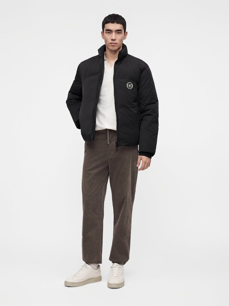 Jeune homme portant une doudoune noire, une chemise blanche, un pantalon marron et des baskets blanches, debout devant un fond neutre.