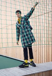 Homme en manteau à carreaux et pantalon noir se tient derrière un filet vert, le tenant d'une main, contre un mur en bois.