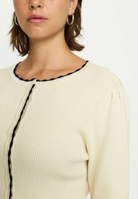 Cremfarbener gerippter Cardigan mit rundem Halsausschnitt, schwarzem gewelltem Rand und Knopfverschluss. Der Puffärmel sorgt für eine interessante Textur im Design.