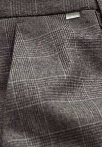 Grauer karierten Stoff mit strukturierter Webart. Verfügt über subtile Linien und verschiedene Schattierungen, die ein strukturiertes Erscheinungsbild erzeugen. Enthält ein kleines Logoetikett.