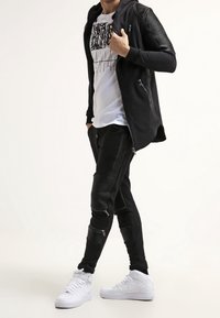 Felpa nera con zip e maniche textured, t-shirt bianca con grafiche, pantaloni jogger neri con cerniere e sneakers bianche.