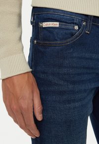 Jeans de mezclilla azul oscuro con costuras visibles, que presentan una pequeña etiqueta roja y blanca de Calvin Klein en el bolsillo delantero y detalles en metal plateado.