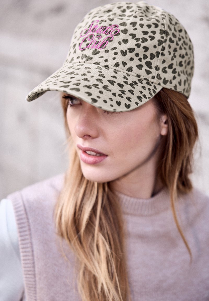 Femme aux longs cheveux blonds portant une casquette beige à motif léopard avec une broderie rose "Swag Club" et un pull sans manches rose clair.