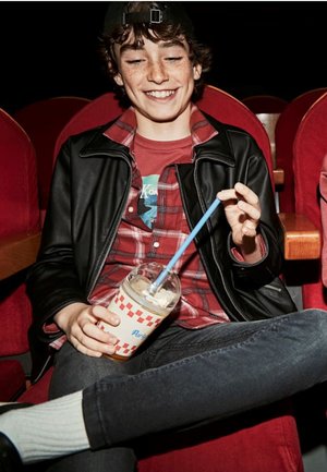 Giacca di pelle nera sopra una camicia di flanella rossa. Tieni un drink con una cannuccia blu, seduto su una poltrona da teatro con tappezzeria rossa e braccioli in legno.