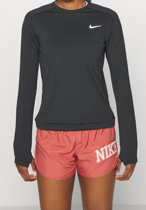 Osoba ubrana w czarną koszulkę Nike z długim rękawem i czerwone spodenki Nike z białym logo na lewym udzie na jednolitym tle.