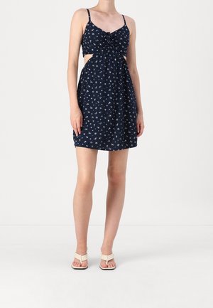 Kobieta ubrana w granatową mini sukienkę bez rękawów z białym kwiatowym wzorem, z wycięciami po bokach, oraz beżowe buty na obcasie z odkrytymi palcami, stojąca na białym tle.