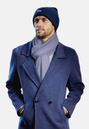 Cappotto navy doppiopetto in misto lana, sciarpa a coste blu chiaro e berretto di maglia blu scuro con una piccola etichetta color sabbia. Progettato per il freddo.