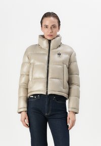 Beige pufferjack met hoge kraag, ritssluiting en een externe pocket; heeft een boxy vorm en glanzende textuur. Gekocht met donkere jeans.