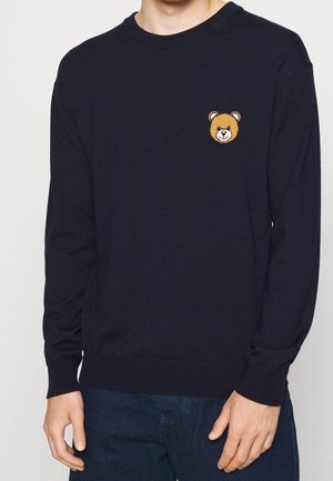 Hombre con suéter de manga larga azul marino con un pequeño emblema de la cara de un osito en el pecho izquierdo, combinado con jeans azul oscuro.