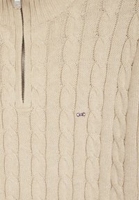 Pull en tricot beige avec un motif en torsades, doté d'un col zippé court et d'un petit logo violet sur le côté gauche.