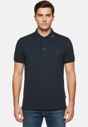 Uomo che indossa una polo nera aderente con maniche corte e tre bottoni, abbinata a jeans blu, in piedi davanti a uno sfondo semplice.