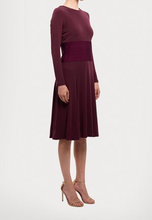 Femme portant une robe longue à manches longues, violet foncé, longueur genou, avec une large ceinture assortie et des sandales à talons hauts couleur nude, debout de profil.