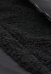 Schwarzes, mit Fleece gefüttertes Jackeninnere, das ein strukturiertes, flauschiges Material und eine glatte Außenseite mit Reißverschlussdetail entlang der Kante zeigt.