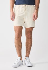 Pantaloni corti beige in cotone con vita elastica e cordoncino. Texture liscia, lunghezza fino a metà coscia, abbinati a sneakers bianche.