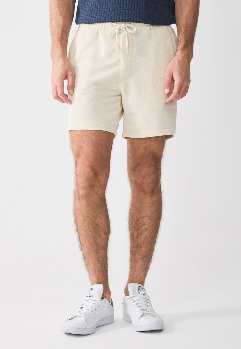Shorts en coton beige avec une taille élastique et un cordon de serrage. Texture lisse, longueur mi-cuisse, associés à des baskets blanches.