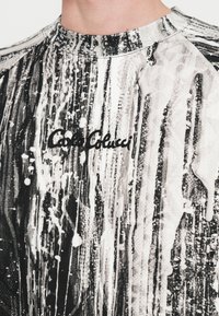 Fekete-fehér grafikus pulóver festékszerű fröccsenésmintával, bordázott, kerek nyakkivágással és fekete hímzett logóval.