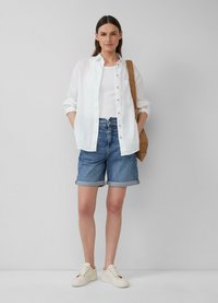 Chemise blanche en lin à boutons sur un débardeur blanc, associée à un short en denim bleu. Pour parfaire le look, une paire de baskets blanches et un sac en osier.