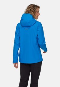 Giacca impermeabile blu con cappuccio, dotata di chiusura zip e polsini elasticizzati. Logo sul retro ben visibile; progettata per attività all'aperto.