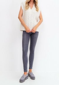 Roomkleurige gebreide mouwloze trui met V-hals en een gestructureerd kabelpatroon, gecombineerd met grijze skinny jeans en grijze plateau loafers.