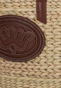 Geweven strooien mand met een rond bruine leren patch met een verhoogd ontwerp en witte stiksels, die de textuur en vakmanschap benadrukt.