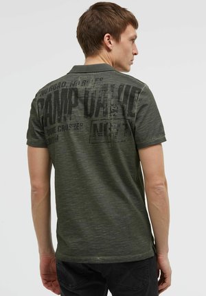 Young man facing away wearing a dark green short-sleeve polo shirt with large black printed text on the back.