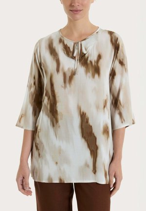 Donna che indossa una maglia larga beige e marrone tie-dye con maniche 3/4 e un piccolo dettaglio annodato sul davanti, abbinata a pantaloni marroni.