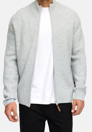 Mann trägt hellgrauen, gerippten Reißverschluss-Pullover über schlichtem weißen T-Shirt und schwarzer Hose vor weißem Hintergrund.