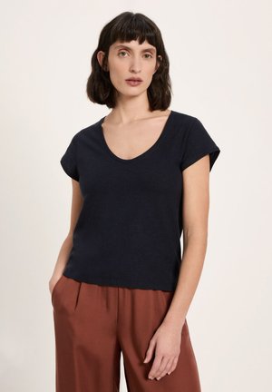 Frau mit kurzen dunklen Haaren trägt ein schwarzes V-Ausschnitt-T-Shirt mit kurzen Ärmeln und rostfarbene, weit geschnittene Hosen, stehend vor einem einfarbigen Hintergrund.