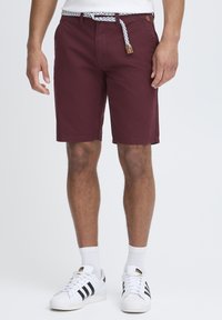 Shorts bordeaux avec une taille texturée, présentant un design en corde tissé et un accent en cuir. Associés à des baskets blanches et des chaussettes mi-mollet.