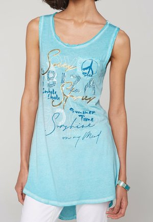 Femme portant un débardeur bleu clair sans manches avec des mots et des motifs imprimés, assorti d'un pantalon blanc et d'un bracelet multicolore.