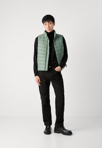Vesta căptușită verde deschis cu un model cu dungi orizontale, purtată peste un pulover cu guler înalt negru și pantaloni negri, asortată cu pantofi negri și o căciulă.