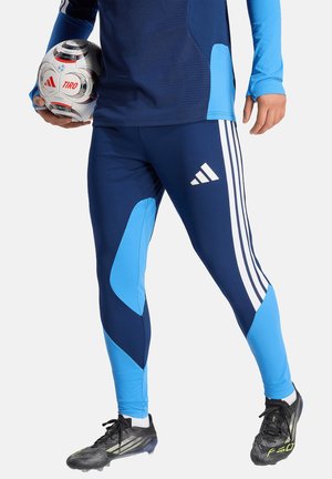Osoba ubrana w niebieską odzież sportową Adidas trzyma białą i czerwoną piłkę piłkarską Adidas Tiro, stojąc na szarym tle w czarnych korkach.