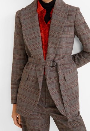 Blazer ceinturé à carreaux gris et marron avec pantalon assorti, porté sur un chemisier en dentelle rouge et un col roulé noir.