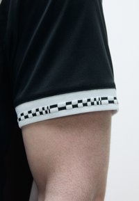 Svart sportsjersey-sleeve med teksturert stoff, med en hvit mansjett prydet med et svart og hvitt rutete mønster.