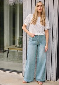 Top blanco de eyelet con mangas abullonadas, jeans de pierna ancha de cintura alta en azul claro y sandalias de tiras. Frente a una pared de madera.