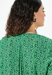 Blusa verde con un design a fantasia con macchie nere, texture morbida e un dettaglio plissettato sul retro.