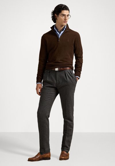 Suéter marrón con cremallera y cuello alto, de mangas largas, sobre una camisa azul de cuello. Pantalones grises con pliegues y zapatos de cuero marrón completan el conjunto. Lleva gafas.