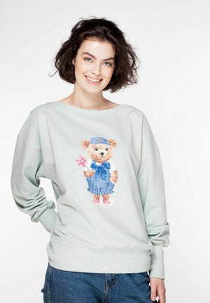 Jasnozielona bluza z nadrukiem niedźwiedzia ubranego w niebieski strój, trzymającego różowy kwiat, z prążkowanymi mankietami i paskiem w talii.
