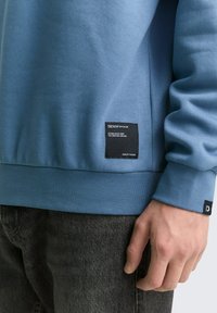 Zila kokvilnas sporta krekls ar ribotajiem piedurknēm, kas iekļauj auduma birku ar zīmola un kvalitātes informāciju. Valkāts virs tumšām džinsa biksēm.