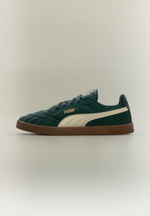 Σκούρο πράσινο αθλητικό παπούτσι Puma με λευκή ρίγα, καφέ σόλα και χρυσό λογότυπο Puma στο πλάι και στη γλώσσα, σε προφίλ σε ανοιχτό φόντο.
