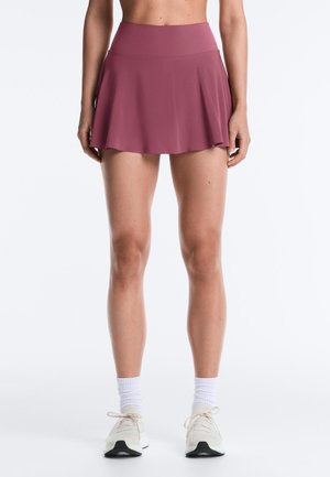 Femme portant une jupe-short de sport mauve, des chaussettes blanches courtes et des chaussures de course écrues, debout devant un fond blanc uni.