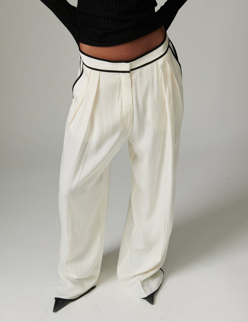 Άτομο που φοράει λευκό παντελόνι υψηλής μέσης και φαρδιάς γραμμής με μαύρη επένδυση, συνδυασμένο με μαύρα μυτερά παπούτσια και κοντό μαύρο πουλόβερ.