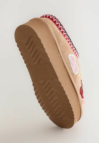 Tan suede shoe with a textured rubber sole. Features red and black decorative stitching and embroidered patches, including a cherry and text.
