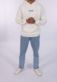 Sudadera crema con texto negro "crystal" y "dreams". Bolsillo frontal, puños acanalados, combinada con vaqueros azul claro y zapatillas blancas.