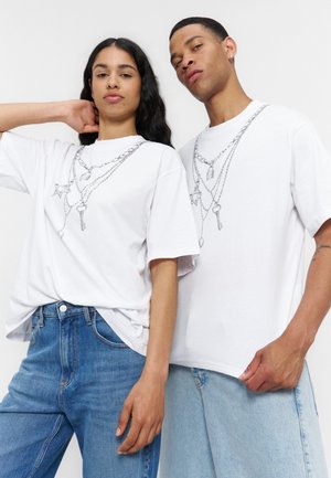 Deux personnes portant des t-shirts graphiques blancs avec des motifs de chaîne et de clé, associés à des jeans en denim bleu, posant devant un fond uni.