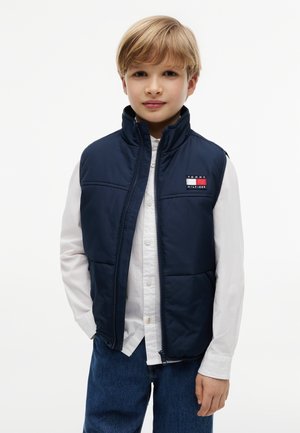 Blonder Junge trägt eine marineblaue gesteppte Weste über einem weißen Hemd mit Knöpfen und blauen Jeans, steht mit den Händen in den Taschen vor weißem Hintergrund.